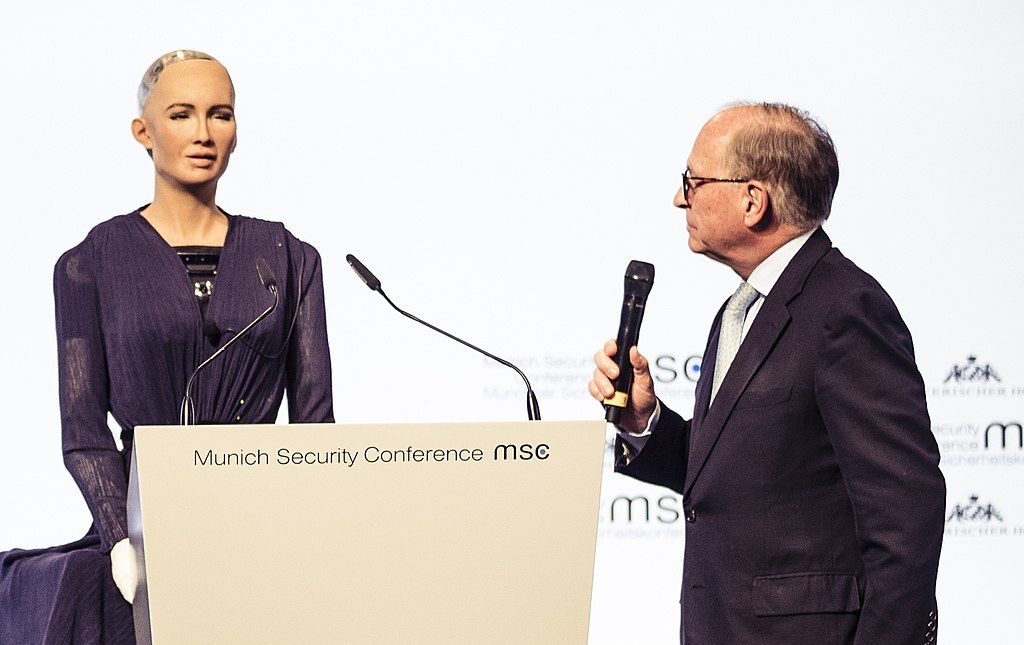 Sophia the AI robot stands at a podium in front of a guest speaker